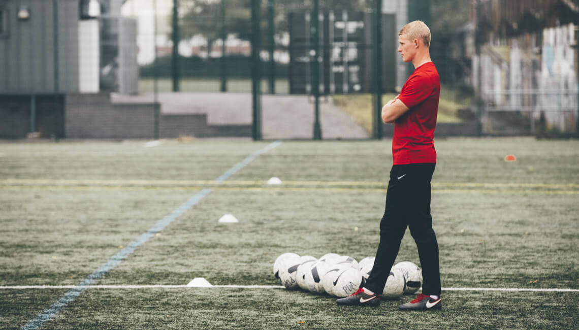 A man in training gear standing with a pile of training footballs